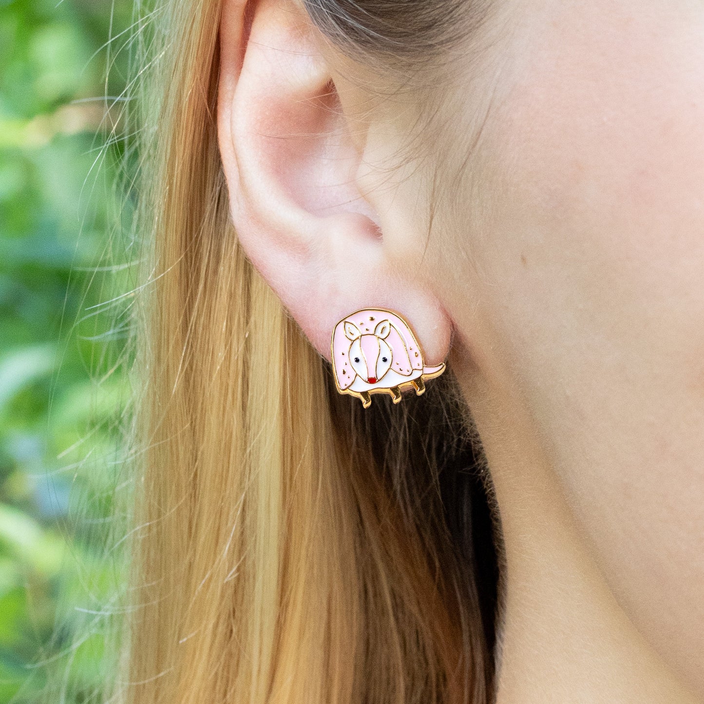 Close-up of an ear wearing a pink armadillo earring with a green outdoor background
