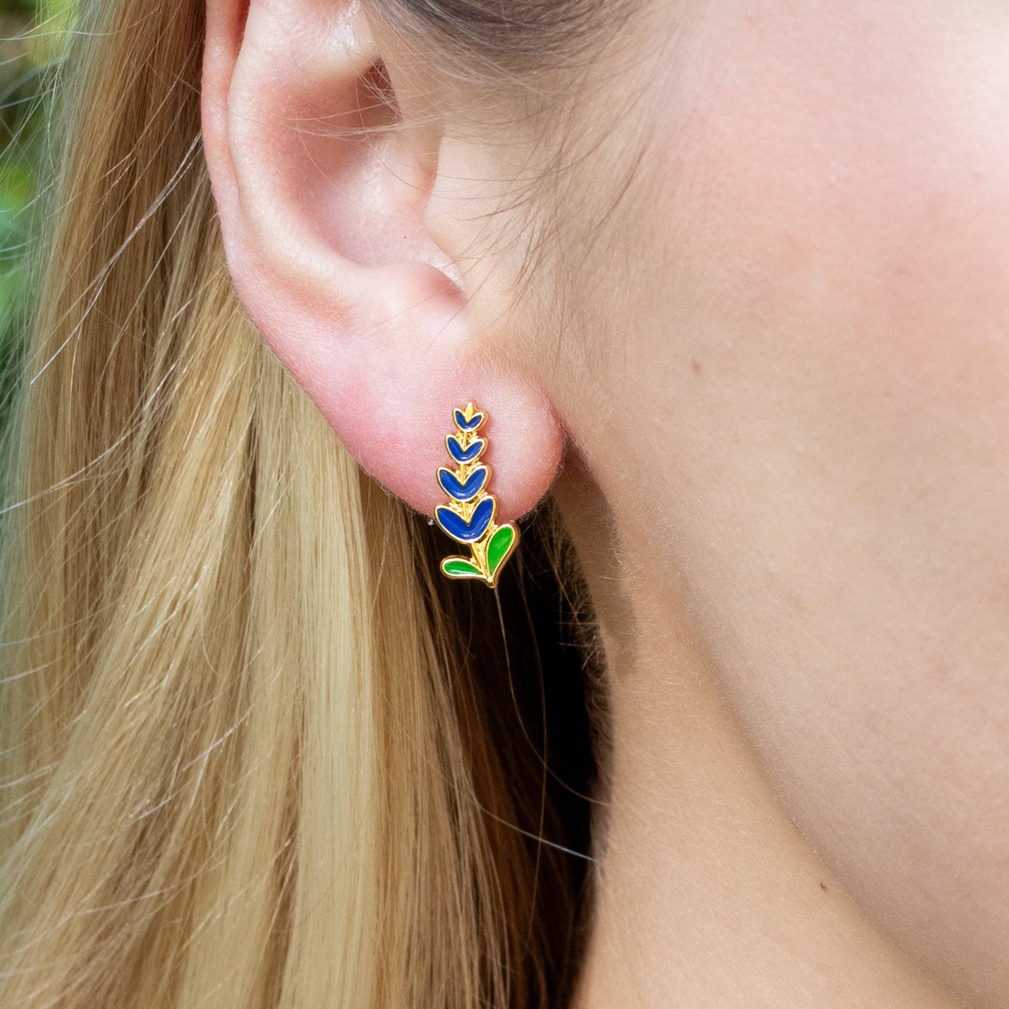 Close-up of an ear wearing Texas bluebonnet earrings with a blurred natural background.