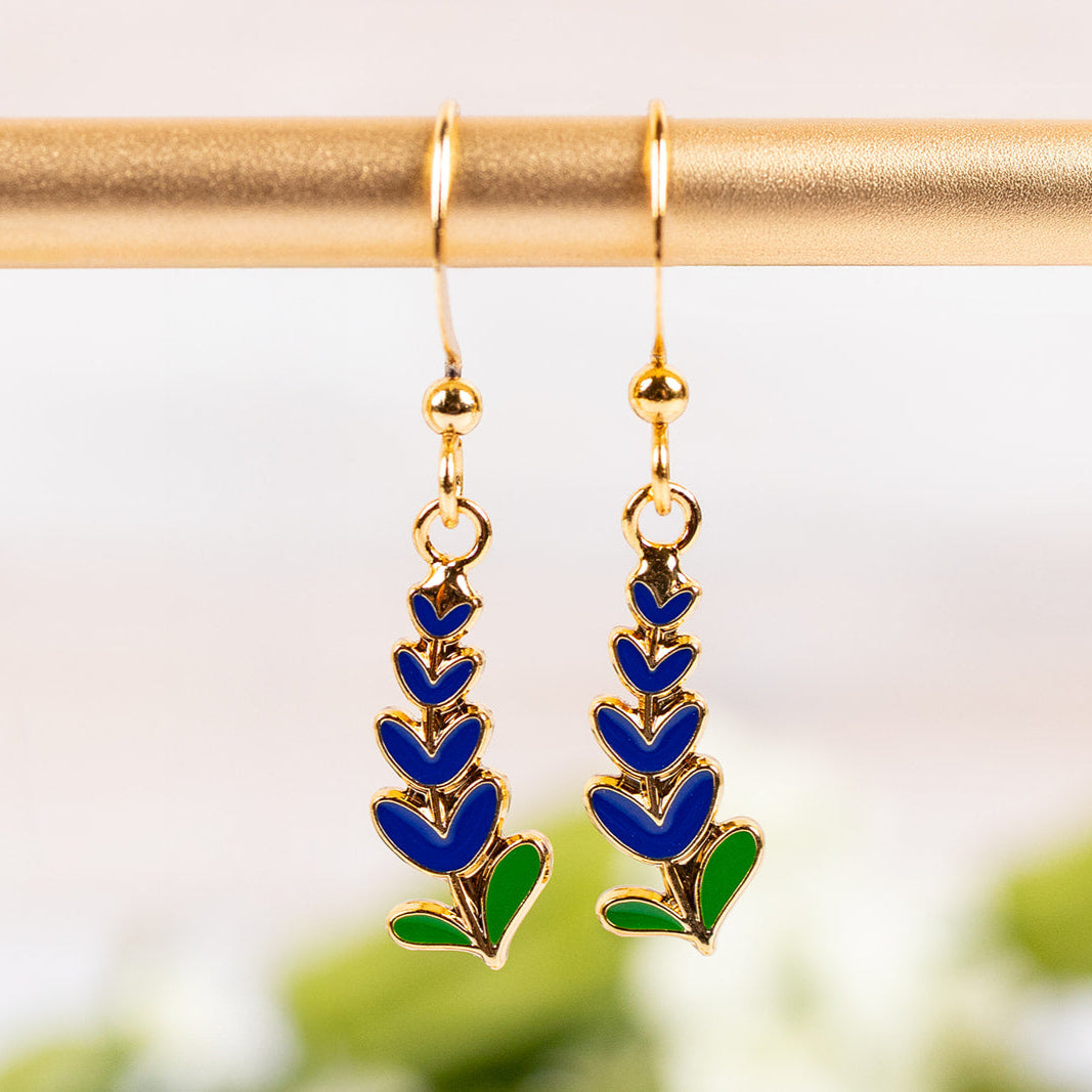 Drop earrings with Texas Bluebonnet designs on a gold rod against a blurred natural background.