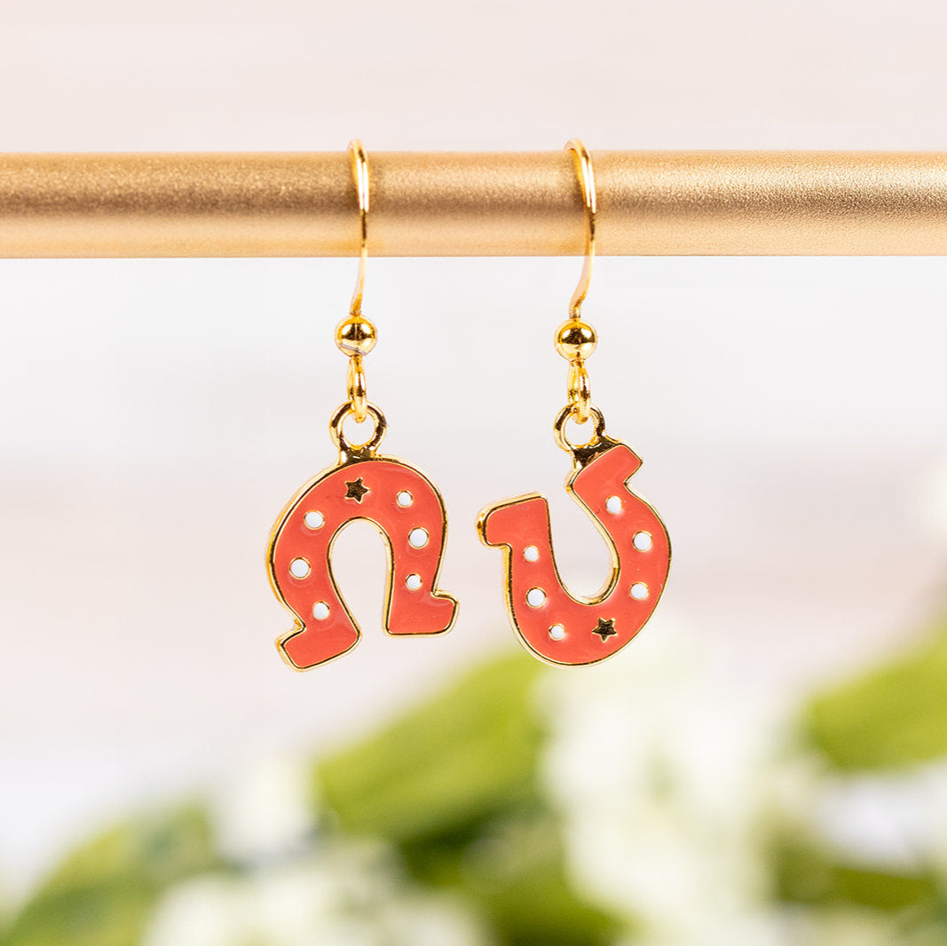 Orange horseshoe-shaped earrings on a gold hook against a blurred green and white background