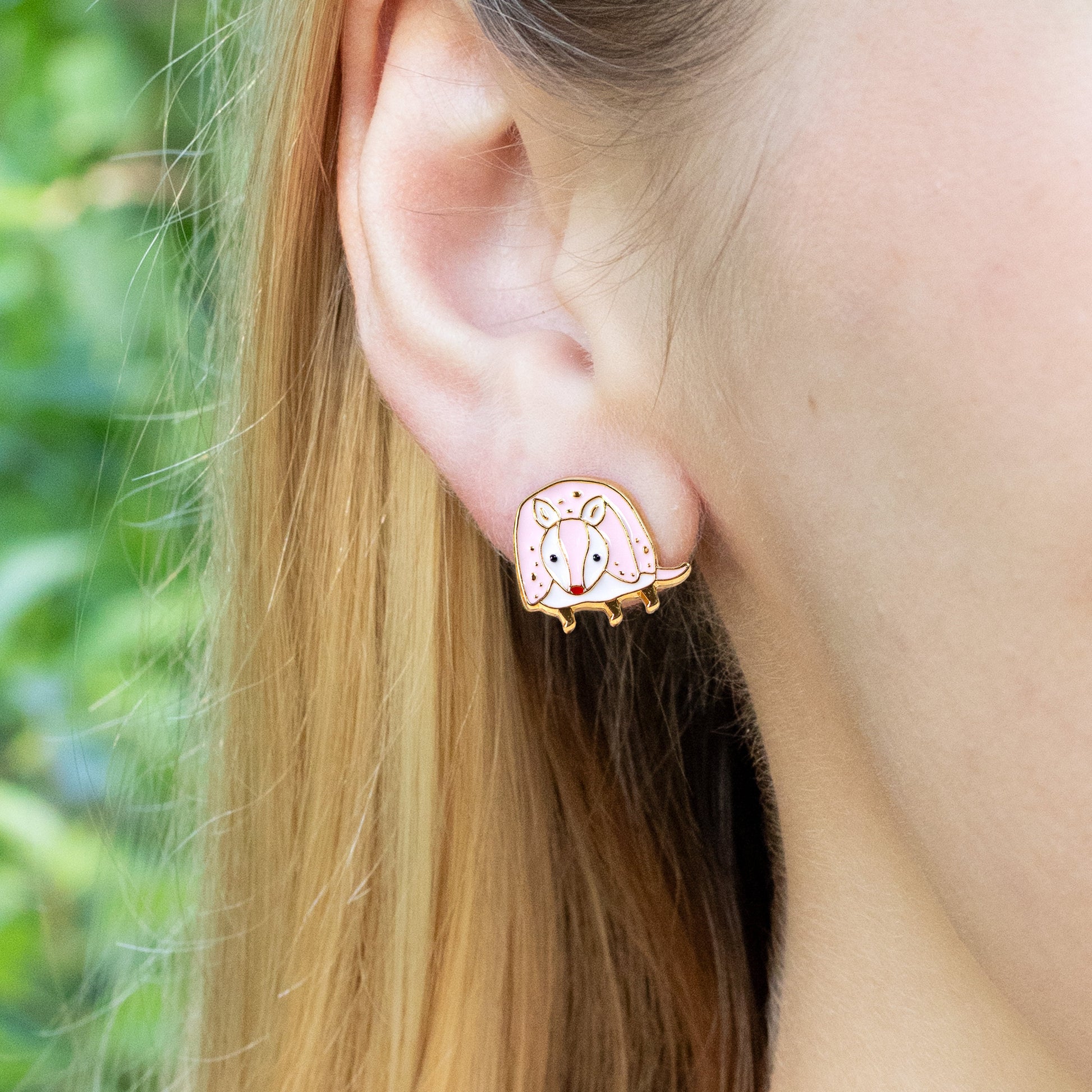 Close-up of an ear wearing a pink armadillo earring with a green outdoor background
